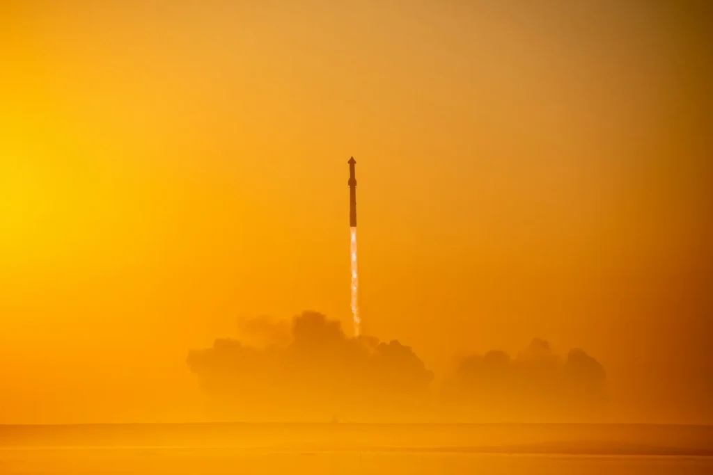 Start rakety na pozadí oranžové oblohy při východu nebo západu slunce, s oblakem kouře pod ní.