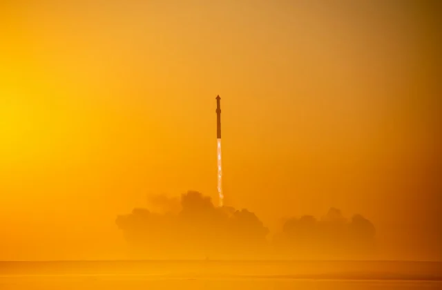 Start rakety na pozadí oranžové oblohy při východu nebo západu slunce, s oblakem kouře pod ní.