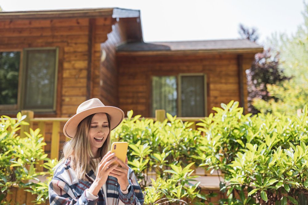 Žena s kloboukem držící mobilní telefon před dřevěným domem, ideální pro práci na dálku s využitím půjčeného Starlink internetu.