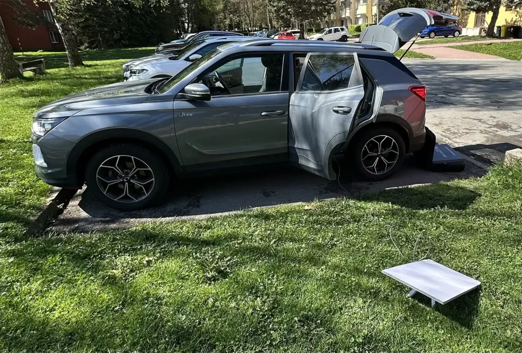 Šedé SUV SsangYong / KGM Korando zaparkované na parkovišti s otevřenými zadními dveřmi a kufrem. Vedle auta na trávě je satelitní anténa Starlink připojená kabelem do vozu.