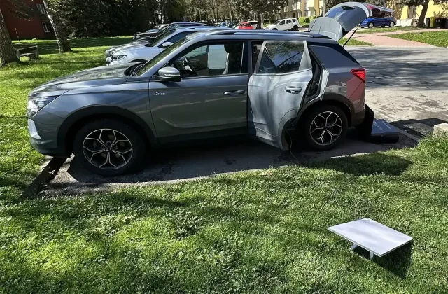 Šedé SUV SsangYong / KGM Korando zaparkované na parkovišti s otevřenými zadními dveřmi a kufrem. Vedle auta na trávě je satelitní anténa Starlink připojená kabelem do vozu.